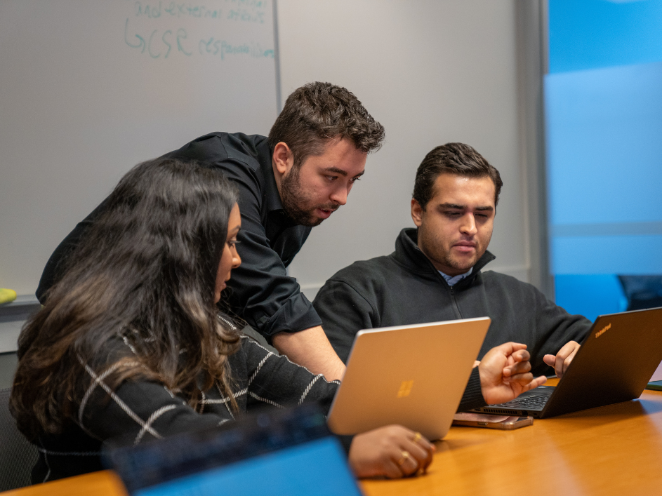 Three MBA students collaborate in a conference room, discussing a laptop's content while one points at the screen.