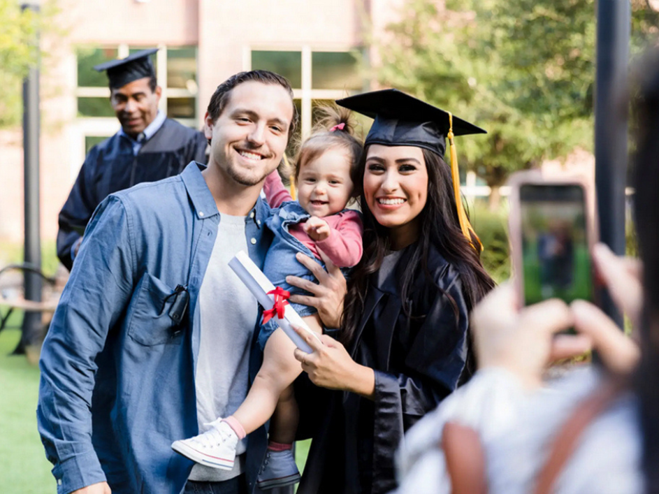 A proud mother in a graduation gown smiles while holding her degree scroll, with her husband and baby beside her.